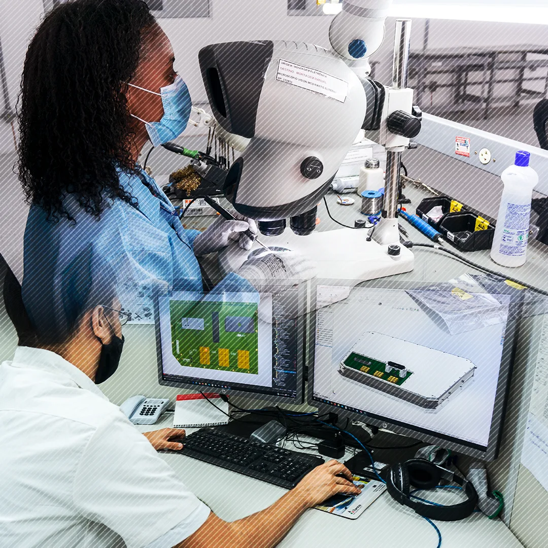 Técnica de eletrônica da IMBEL realizando montagem de placa sob microscópio, com layout e modelo 3D da placa nas telas ao lado